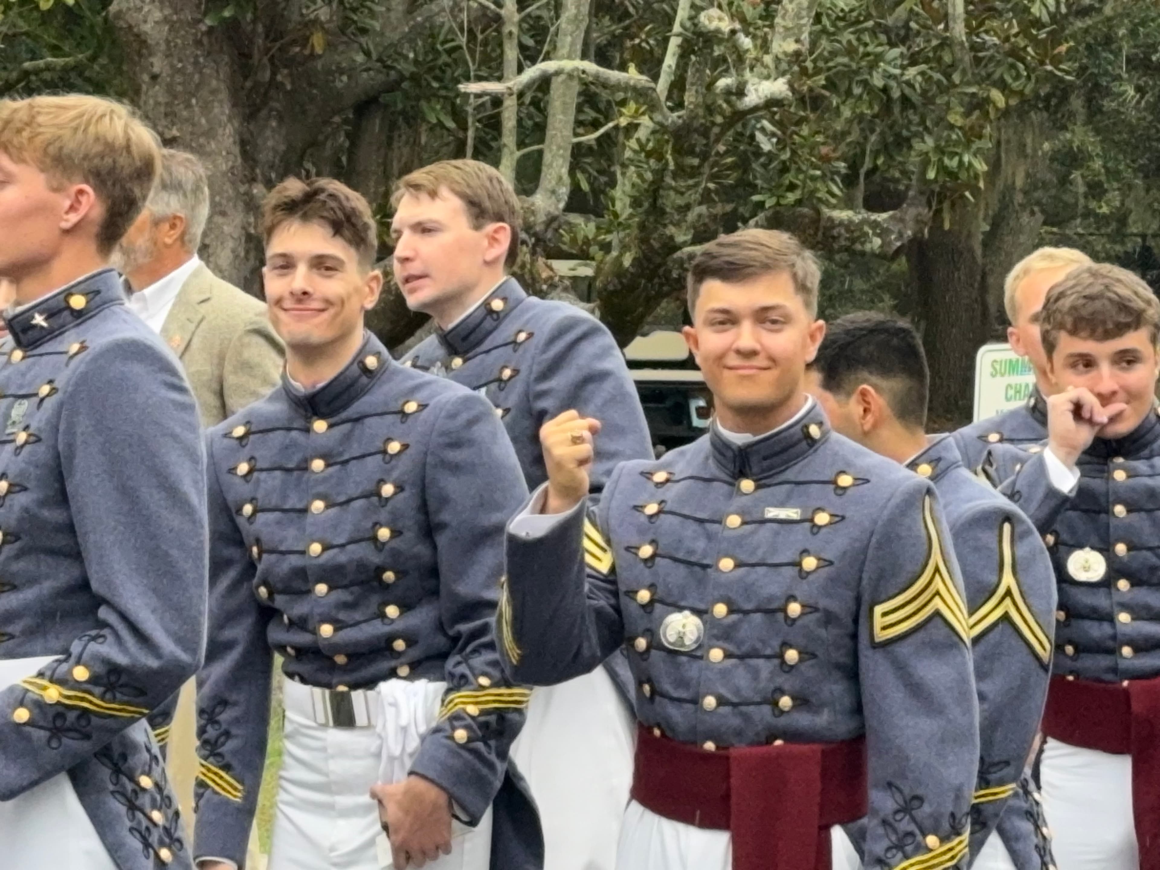 Cadets in dress gray uniforms stand together beneath live oaks on The Citadel campus.