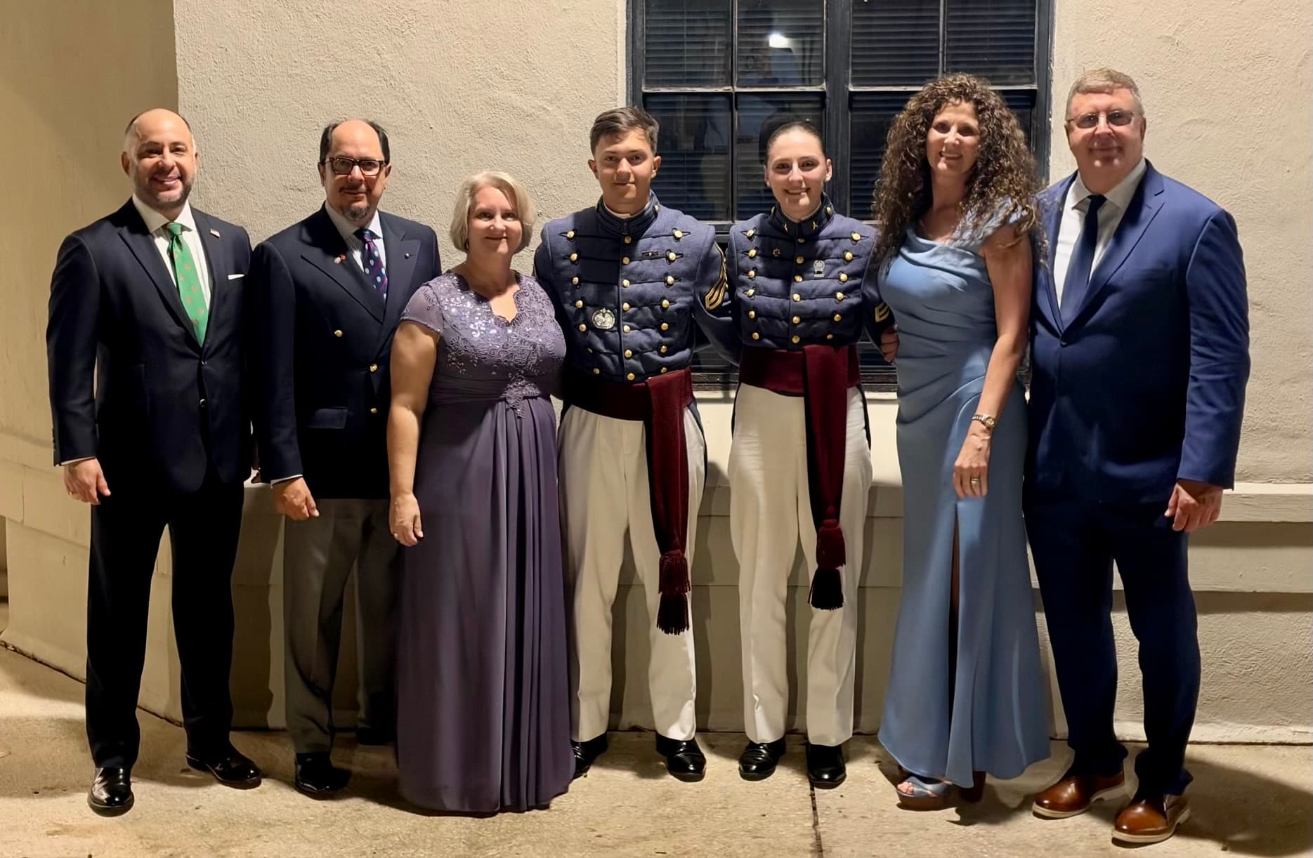 Two cadets in dress gray uniforms with maroon class sashes pose with family in formal attire outside at The Citadel in the evening.